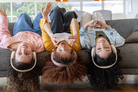 Upside-down teenage friends listening to music on sofa wearing headphones and smiling