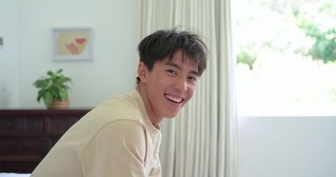 Smiling young asian man sitting on bed by window in bright minimalist bedroom