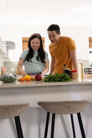 Asian couple preparing healthy meal at bright modern kitchen island, chopping herbs