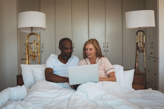 Senior couple in pajamas smiling and using laptop together in bed. Perfect for illustrating themes of senior life, diversity, technology use among the elderly, and home quarantine during lockdown. Suitable for articles on relationships, health and wellness, and remote connectivity.