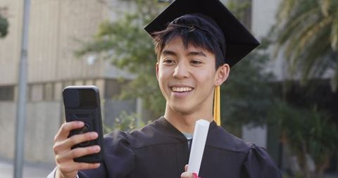 Asian college graduate smiling and taking selfie on campus holding diploma in cap and gown