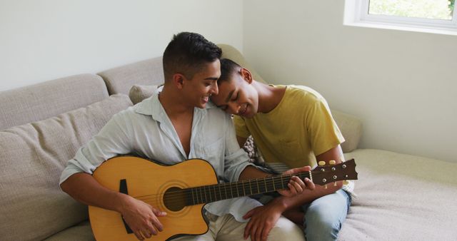 Smiling biracial gay male couple sitting on sofa and embracing while one plays guitar. staying at home in isolation during quarantine lockdown.