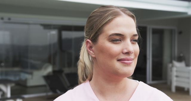 Woman is standing outside a modern home, wearing a pink shirt and smiling. Her surroundings reflect a peaceful and relaxing environment. Perfect for ads and articles related to health, well-being, modern living, and lifestyle blogs.