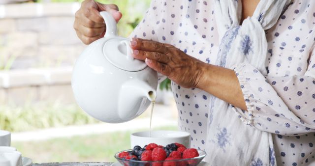 Person pouring tea from a white teapot into a bowl of fresh mixed berries, suitable for use in lifestyle blogs, healthy living articles, outdoor dining features, and casual backyard gathering promotions.
