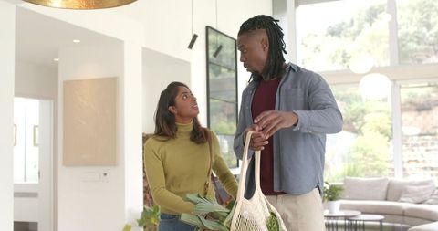 African american man and indian woman carrying market bag of greens in sunlit living room