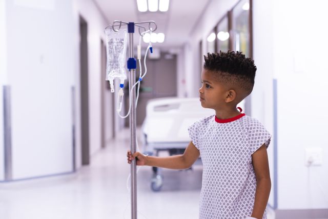 African american boy patient holding drip stand standing in hospital corridor, with copy space. Medical services, hospital and healthcare concept.
