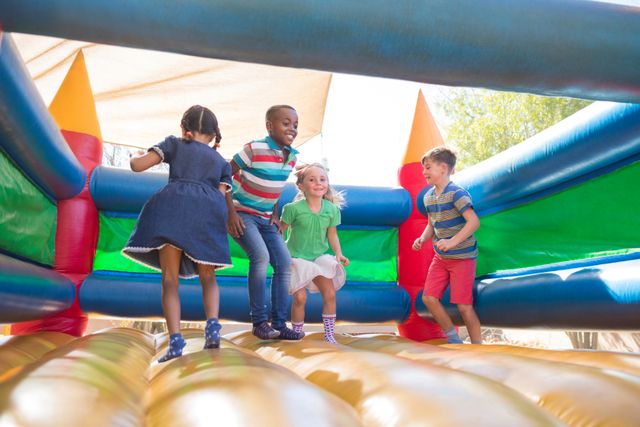 Children are jumping and having fun on a colorful bouncy castle at an outdoor playground. This image is perfect for illustrating concepts of childhood joy, outdoor activities, and group play. It can be used in advertisements for playgrounds, summer camps, or children's events, as well as in educational materials about physical activity and social interaction.