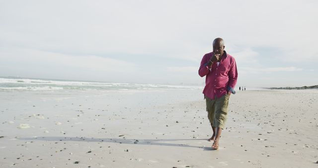 Man enjoying leisurely barefoot walk on sandy beach with calm ocean waves, dressed in casual red shirt and cargo shorts. Ideal for themes about relaxation, leisure activities, vacation, solitude, and nature.