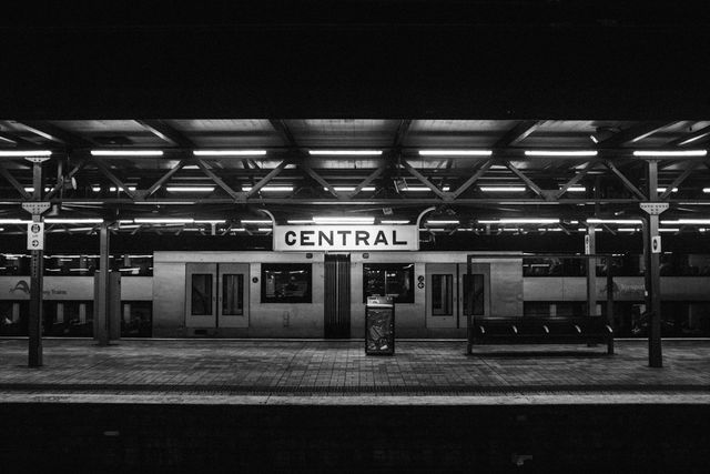 A black and white view of an empty railway station platform at night with a central sign. The photo evokes a sense of solitude and quietness and can be used in materials relating to urban life, transportation, or travel. Suitable for articles, websites, and promotions that want to emphasize calm or deserted urban scenarios.