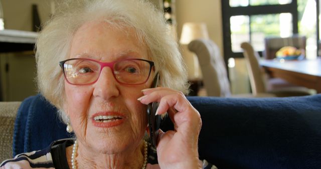 Elderly woman wearing pink glasses is speaking on a smartphone, sitting on a couch in a warmly lit home interior. Suitable for depicting themes related to senior communication, technology adoption by the elderly, staying connected, family relationships, and inclusive interacting. Can be used in health care, family care, senior living, and technology-related advertising and marketing materials.