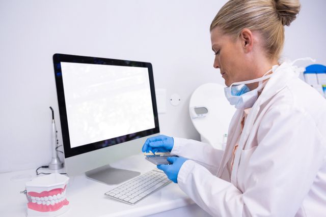Dentist wearing gloves and lab coat working on dental model while sitting by computer in modern clinic. Ideal for use in healthcare, dental care, and medical professional contexts, showcasing dental practices and procedures.