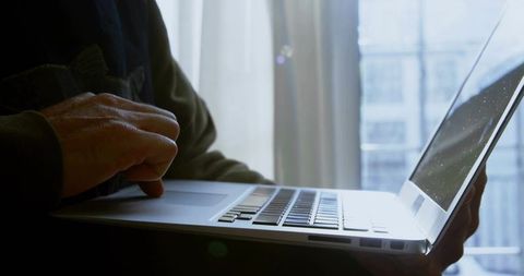 Mature woman typing on silver laptop at window, backlit minimalist urban remote workspace