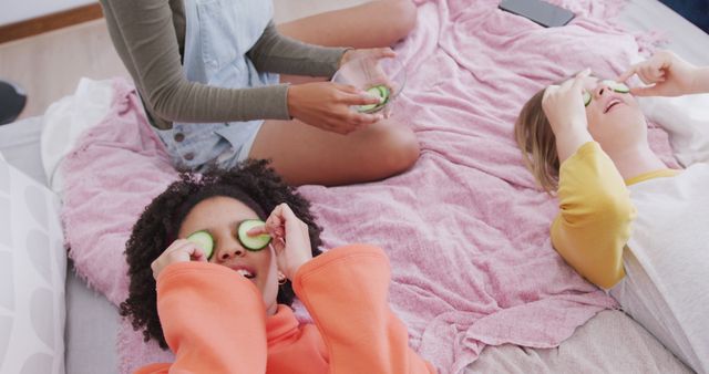 Happy diverse teenager girls lying on bed with cucumber slices on eyes in bedroom. Spending quality time, lifestyle, friendship and adolescence concept.