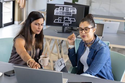 Female colleagues collaborating over laptop while reviewing architectural floor plans in modern work