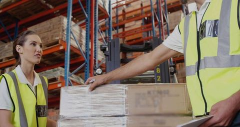 Warehouse workers steadying shrink-wrapped pallet while checking inventory with clipboard