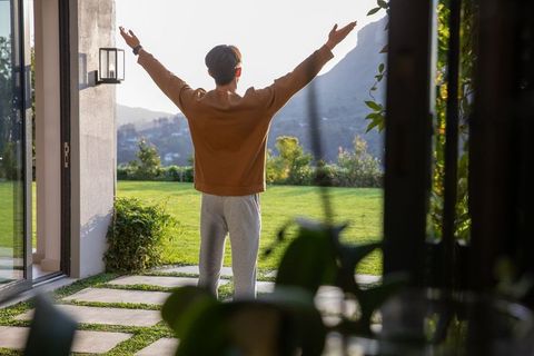 Young man stretching in doorway facing sunlit lawn and mountain view during golden hour