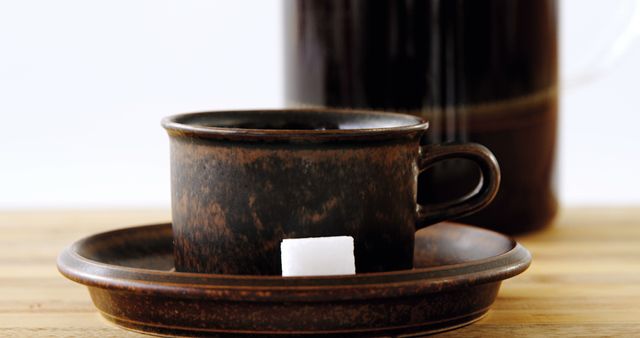 A rustic brown coffee cup sits on a saucer with a single sugar cube beside it, with copy space. The warm tones and simple setup suggest a cozy, inviting atmosphere for a coffee break.