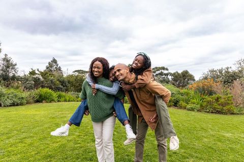 African american family walking in park carrying teenage daughters on parents' backs, smiling