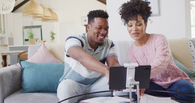 African American couple sitting on sofa using tablet to manage their finances. They are both smiling and seem engaged in the task. Used for themes of financial planning, technology in daily life, teamwork, and modern home living.