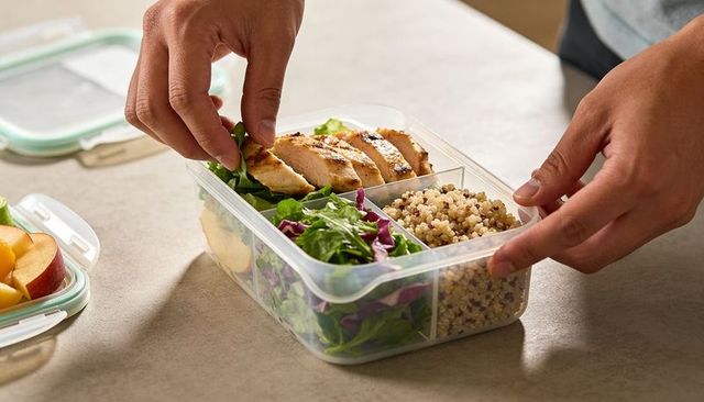 Hands assembling grilled chicken, quinoa and mixed greens lunch box for meal prep