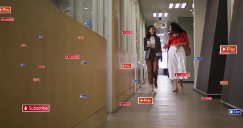 Two businesswomen walking and chatting in modern office corridor holding coffee and phone
