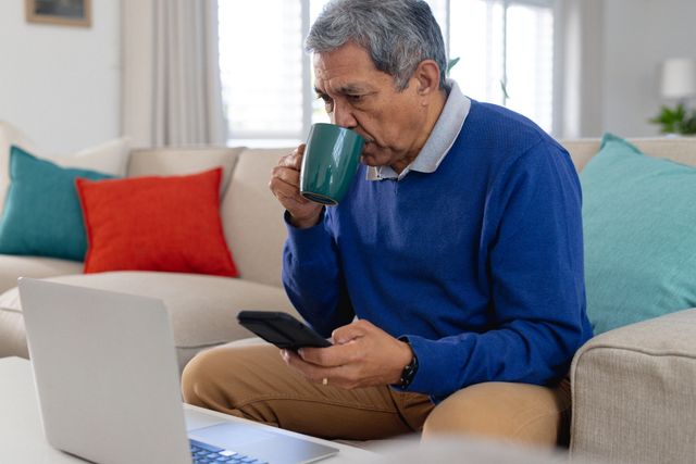 Senior man sitting on couch in living room, using laptop and smartphone while drinking coffee. Ideal for content related to remote work, senior lifestyle, technology use among older adults, and home office setups.