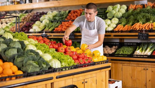 Grocery clerk arranging bell peppers on wooden produce display in bright market aisle