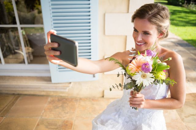 High angle view of happy bride holding bouquet taking selfie with mobile phone while sitting on chair in yard