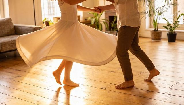 Couple spinning barefoot on sunlit hardwood floor, flared white dress twirling