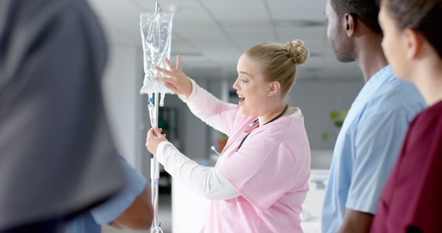 Group of medical professionals in a well-lit hospital environment. Focus is on the blond nurse in pink scrubs explaining the intravenous drip procedure to colleagues. Ideal for use in healthcare training materials, medical education platforms, or hospital promotional content.