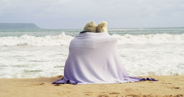 A couple enjoys a serene beach setting, wrapped together in a blanket with a view of the waves, with copy space. Their shared moment by the sea suggests a romantic or peaceful getaway.