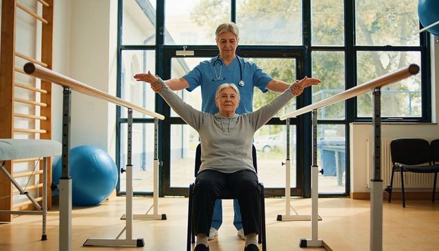 Senior woman raising arms during seated rehabilitation guided by clinician at parallel bars