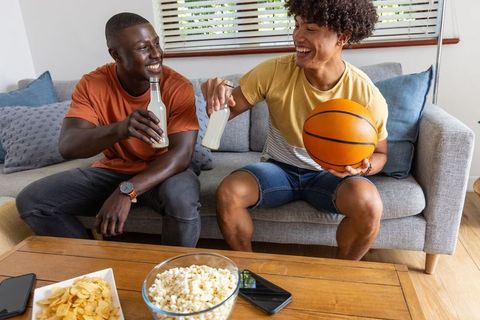 Friends clinking bottles on couch holding basketball with snacks on coffee table