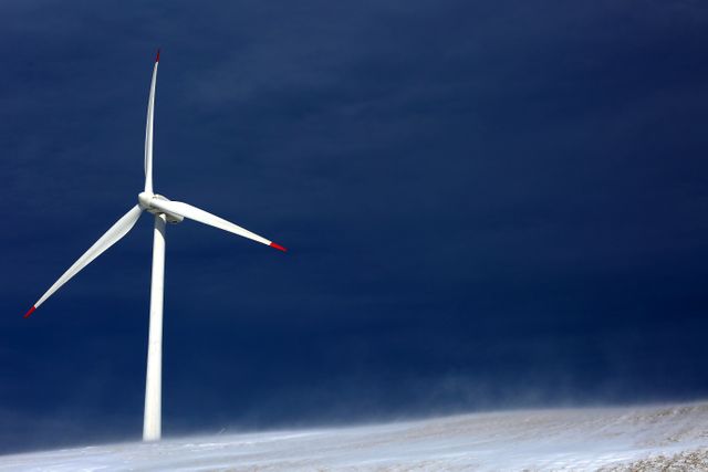Wind turbine shown standing in a snow-covered landscape with a dark clouded sky background. Ideal for visuals related to renewable energy, clean energy sources, environmental conservation, and winter scenes. Suitable for use in environmental campaigns, articles on green energy, climate change, and science publications.