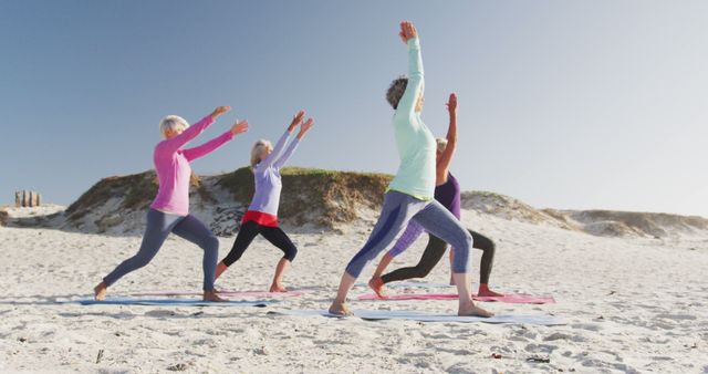 Four senior women practicing yoga on a sandy beach during sunrise. They are doing the warrior pose, wearing athletic clothes. The beach setting with clear sky adds to calm and tranquility. Ideal for themes focused on senior fitness, active aging, wellness, healthy living, retirement activities, and outdoor exercises.
