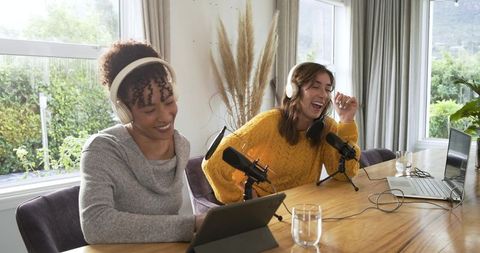 Female cohosts recording home podcast laughing with headphones and pro microphones