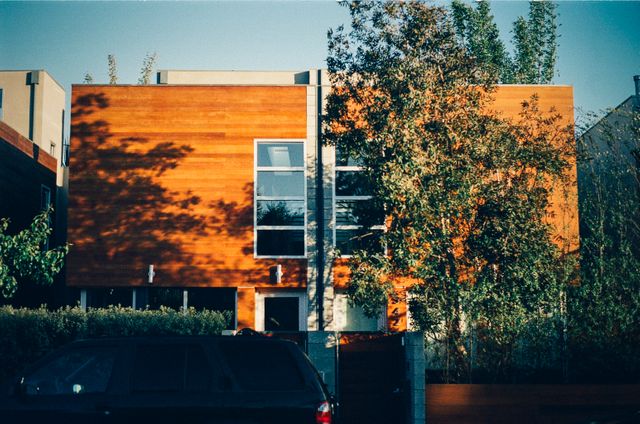 Showcasing a modern house with a sleek wooden facade and large windows surrounded by trees. A black car is parked in front. Ideal for magazines, blogs, and websites focused on architecture, urban living, and property investment.