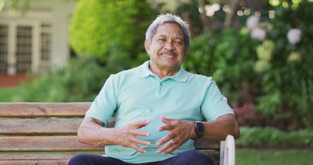 Happy senior man in a casual light blue polo shirt sitting on a wooden bench in a lush park, smiling and enjoying his time. Suitable for themes of retirement, healthy lifestyle, outdoor relaxation, enjoying nature, and elderly well-being in various wellness, healthcare, and lifestyle contexts.