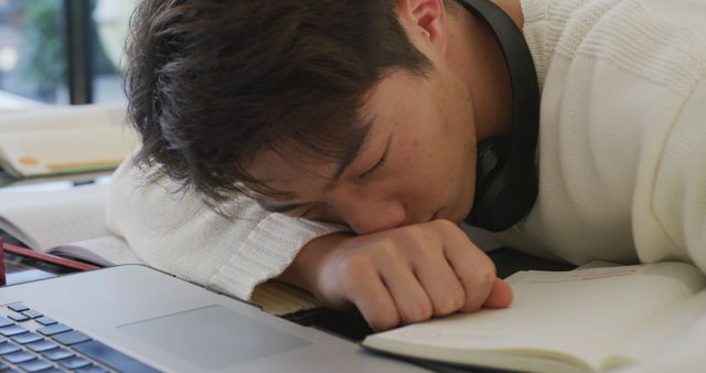 Young man is resting his head on his arms at a desk with an open laptop and notebook. He looks tired from studying or working for a long time and appears to be catching up on sleep. This can be used to depict academic life, work fatigue, or the need for rest. Ideal for educational articles, blogs on work-life balance, or sleep health.
