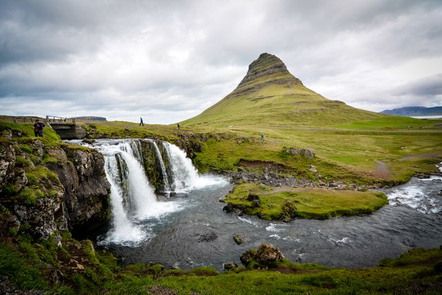 This picturesque landscape features the iconic Kirkjufell Mountain with adjacent waterfalls and flowing river. The overcast sky enhances the striking green grass and rugged scenery, making it an ideal image for promoting travel, adventure tourism, nature appreciation, and outdoor activities. Additionally, it can be used in travel magazines, blogs, and guides for Iceland.