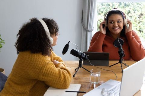 Female podcast hosts recording at home table wearing headphones using microphones