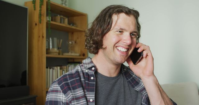 Cheerful man in plaid shirt talking on phone, displaying happiness and engagement. Perfect for illustrating casual communication, remote relationships, indoor activities, telecommuting, or advertising telecom services.