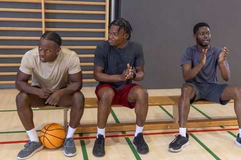 African american basketball players sitting on bench clapping during gym practice
