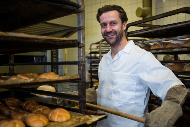 Smiling baker arranging freshly baked buns on a rack in a commercial kitchen. Ideal for use in articles or advertisements related to baking, culinary arts, professional chefs, bakery businesses, and food industry promotions.
