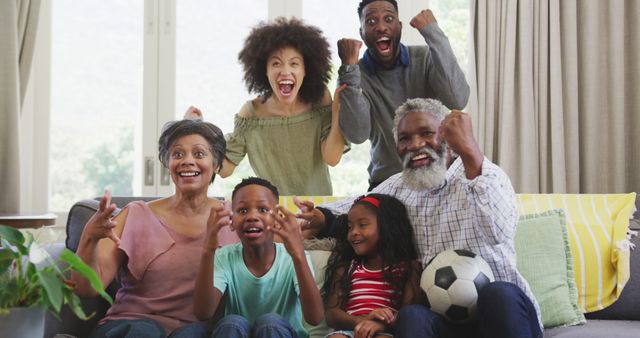 Multigenerational family gathering in living room, showing excitement while watching sports game on couch. Suitable for advertisements or articles about family bonding, sports enthusiasm, home entertainment, or multicultural family experiences.