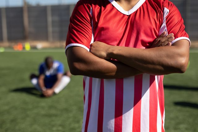 Mid section of biracial male five a side football player wearing a team strip training at a sports field in the sun, standing with arms crossed.