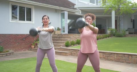Diverse women swinging kettlebells on backyard lawn near patio for outdoor fitness
