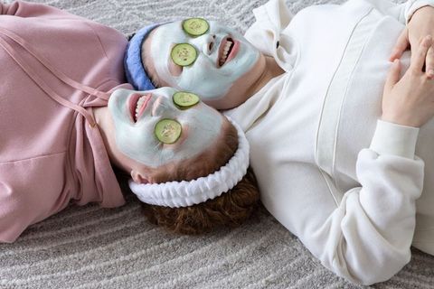 Friends enjoying cozy at-home spa day with facial masks, cucumber slices, laughing together