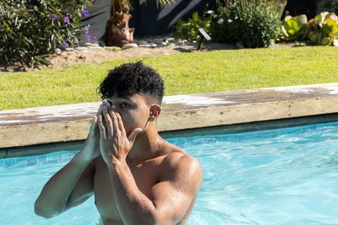 African american man standing chest-deep in backyard pool covering face with hands