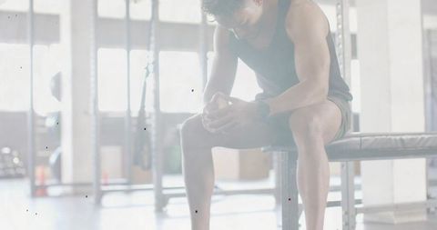 Male athlete resting between sets on bench, leaning forward and clasping hands in gym